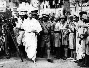 Subhas Chandra Bose wearing all white and a boys cap walks in front is escorted by military soldiers. 