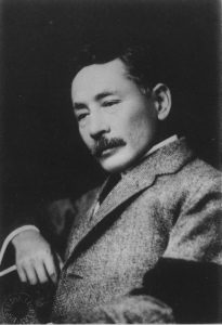 A photograph of middle-aged Natsume Sōseki, distinguished by his short mustache. He is dressed in a Western-style blazer, and in his hand, he holds a pencil, suggesting his engagement in literary pursuits or writing. The image captures the intellectual presence and iconic appearance of Natsume Sōseki, a renowned Japanese author and literary figure.