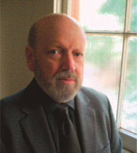 Photograph of a middle aged Damon Wood. He has a bald head and a grey short beard, and he is wearing a black business suit. 