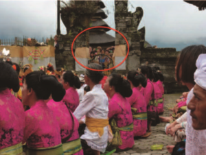A group of villagers sit in a row at a temple in Bali. The women are all wearing pink sarongs with a green sash, while the men wear a white sarong with a yellow sash. They are engaged in a prayer. 