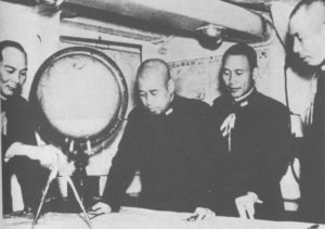 Four Japanese middle-aged men in military uniforms studying a map while gathered around a table. One man holds an army navigator protractor, while a globe sits on the table in front of them. The men appear engaged in a strategic discussion or planning session.