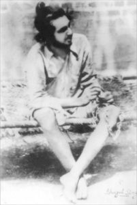 Photograph of Bhagat Singh as a young man. He is sitting on a mat and is wearing a t-shirt and shorts. 